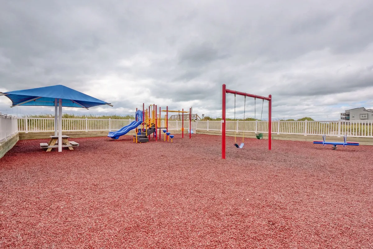 A vibrant playground with colorful climbing equipment and swings, situated next to a picnic area with a large umbrella. The space is alive with children playing and families gathering, evoking a joyful, community-oriented atmosphere.