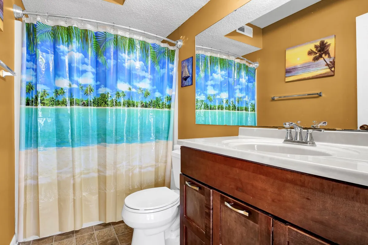 A bathroom featuring a beach-themed shower curtain, a wooden vanity, and decorative wall art, evoking a tranquil, vacation-like atmosphere. The design is clean and functional, offering a refreshing retreat after beach-filled days.