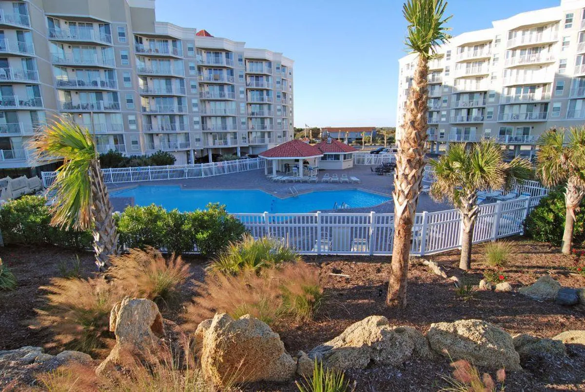 A crystal-clear swimming pool surrounded by palm trees and landscaped gardens, set beside attractive resort buildings. There are lounge chairs and a gazebo, creating an inviting space for swimming, sunbathing, and socializing outdoors.