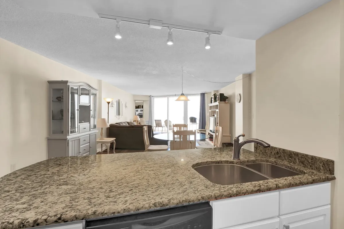 A view from the kitchen showcasing a granite countertop, an adjacent dining area with a table and chairs, and a comfortable living room beyond. Large windows bring in light, and the setup is clearly designed for group meals and social gatherings.