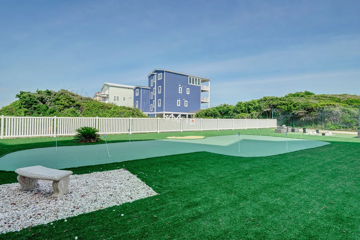 This image features a beautifully maintained artificial putting green surrounded by a landscaped garden, a stone bench for relaxing, and the blue exterior of a house in the background. The scene evokes a sense of outdoor leisure and casual recreation in a tranquil, green setting.