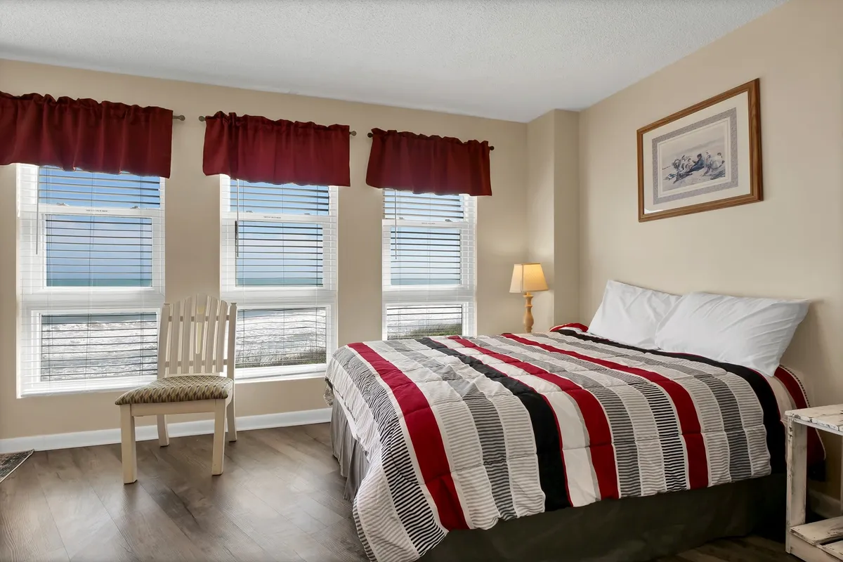 A warm and inviting queen guest room with a striped bedspread, three large windows with blinds, a bedside lamp, framed artwork, and a wooden chair. The room feels bright and comfortable, perfect for guests looking for a peaceful retreat.