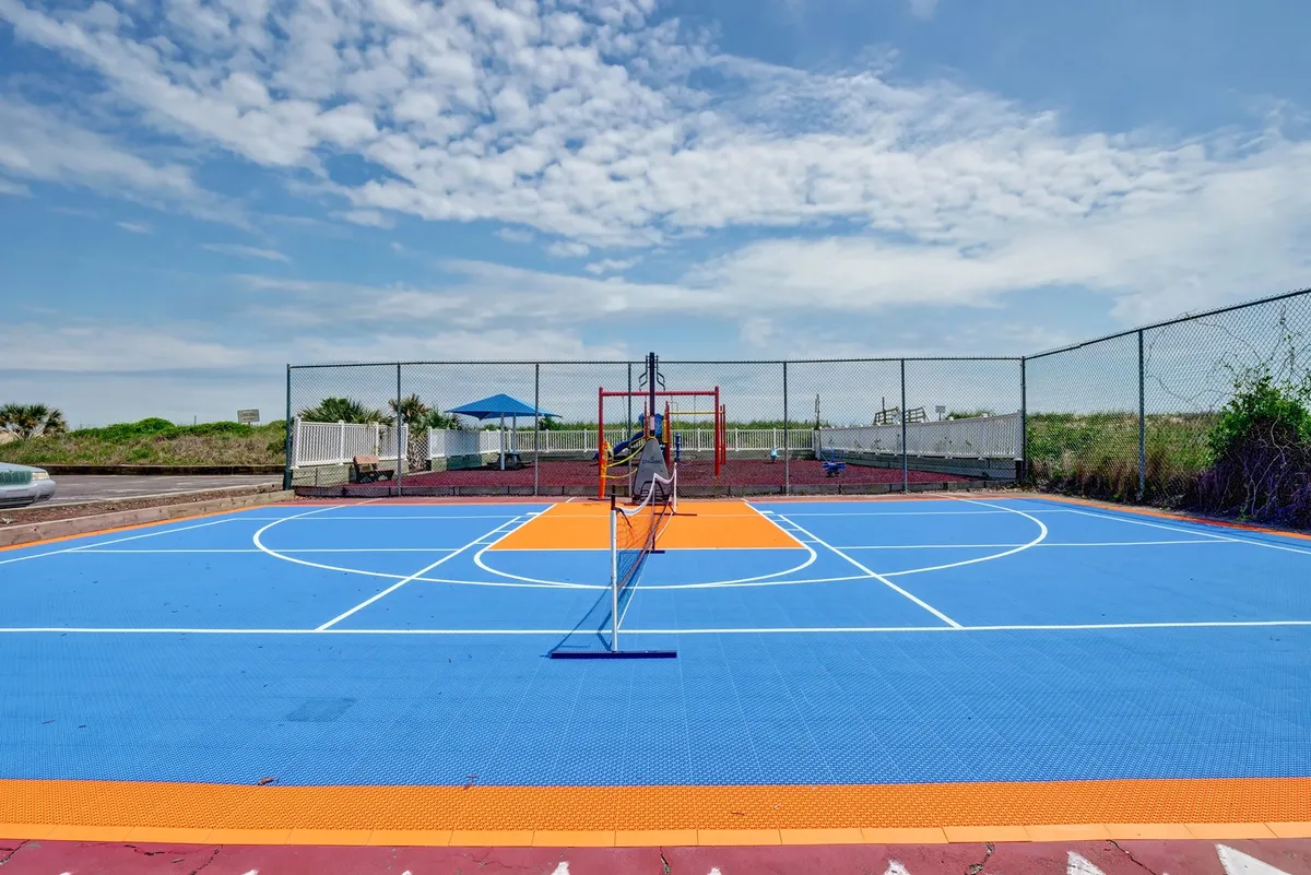 A vibrant outdoor sports area featuring a colorful court suitable for pickleball, tennis, or basketball, surrounded by greenery with a play area in the background. The scene is lively and inviting for group activities and friendly competitions.