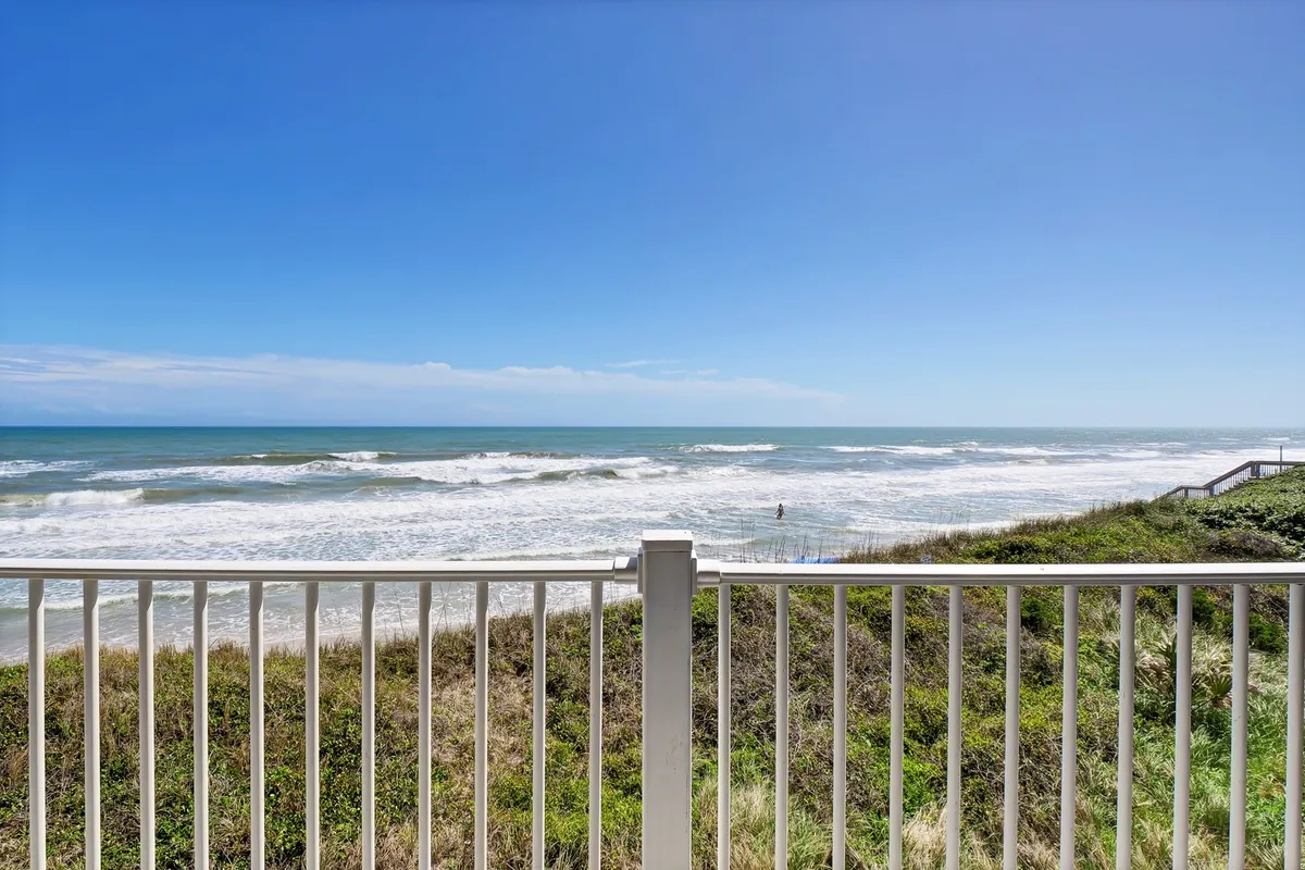 A beautiful view of the Atlantic Ocean, sandy beach, and clear blue sky as seen from behind a balcony railing. The perspective puts viewers in the position of enjoying the scenery directly from a private oceanfront balcony.