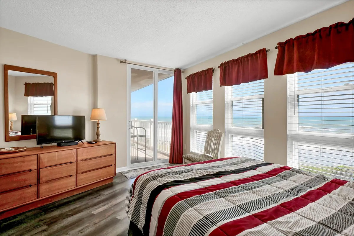 A serene bedroom with a bed dressed in striped bedding, large windows framing an ocean view, a wooden dresser with a TV, and a cozy chair near the window. The space is flooded with natural light and exudes calmness, making it ideal for relaxation and unwinding.