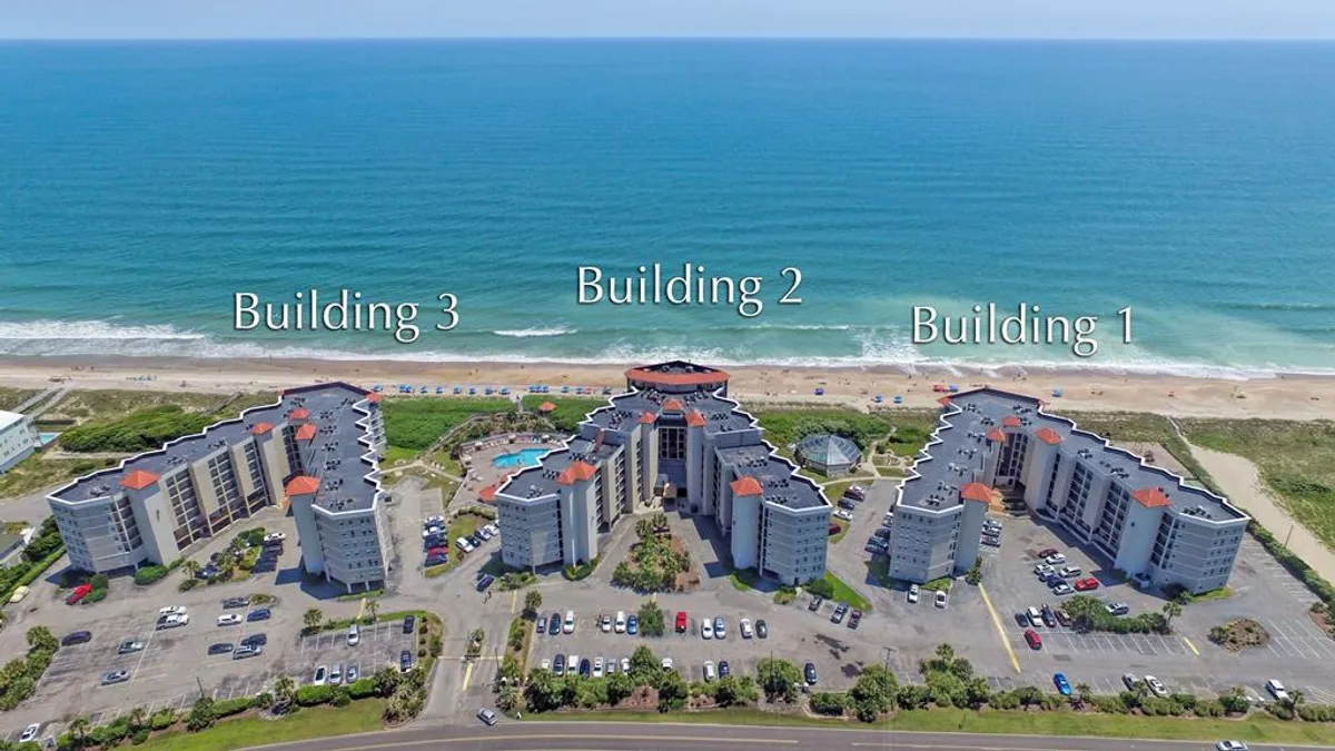 A wide exterior view of the resort complex, featuring three buildings, a sparkling swimming pool, and a sweeping ocean view beyond. The scene combines the resort’s facilities with its prime beachfront location, emphasizing both relaxation and scenic beauty.