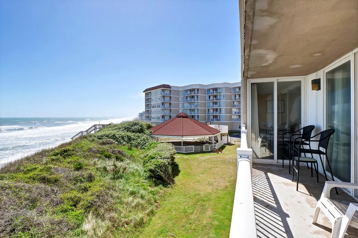 A tranquil oceanfront balcony scene with comfortable seating, lush landscaped gardens below, and sweeping views of the ocean and nearby buildings. The space suggests a perfect setting for group relaxation, morning coffees, or evening chats with family against a coastal backdrop.