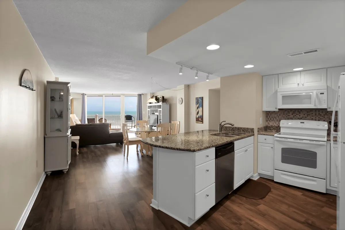 A spacious kitchen with a central island and ocean views visible through large windows. The living area with cozy sofa seating is adjacent, creating an open-concept space ideal for cooking, entertaining, and relaxing. The kitchen features modern appliances and ample counter space, while the windows fill the area with natural light, making it the heart of the home.