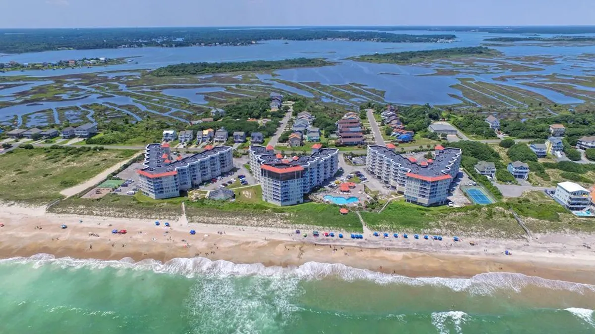 An inviting beachfront property featuring a sparkling swimming pool sits adjacent to soft marshlands and sandy shores. Colorful umbrellas dot the beach beyond, while the poolside area offers spaces for swimming and lounging. The scene captures the seamless connection between the resort, the pool, and the ocean just steps away.
