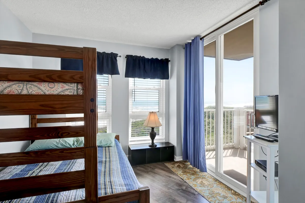 A comfortable, family-friendly bunk room featuring a sturdy bunk bed, large windows, a lamp, and a television. The space is brightly lit with natural light, making it suitable for children or friends to share stories, read, or enjoy a movie night together.