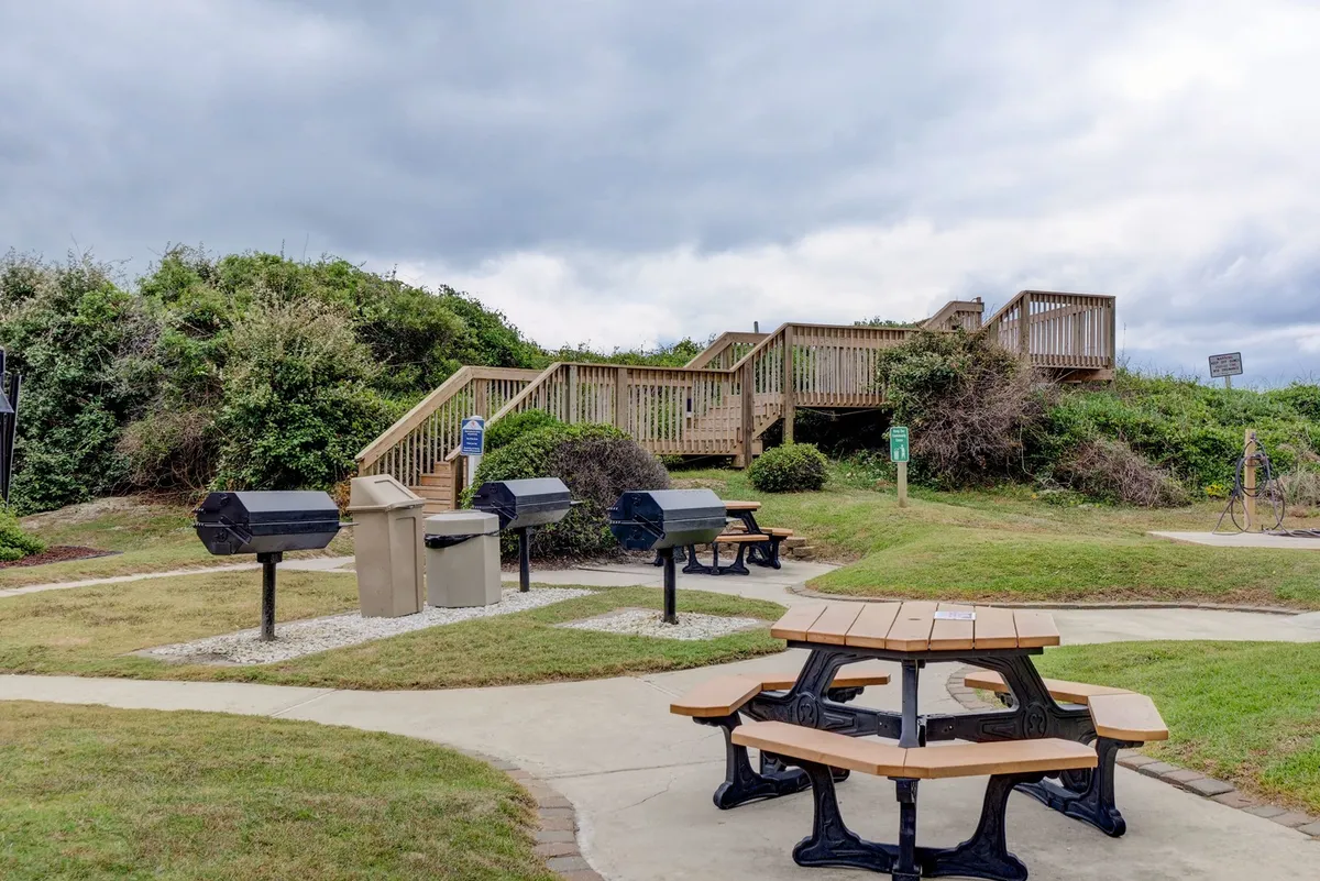 A relaxed communal outdoor grilling and picnic area featuring sturdy picnic tables, multiple grills, and a staircase. The space is designed for barbecues, group meals, and socializing in a casual outdoor setting.