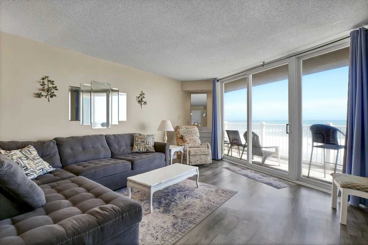 A large sectional sofa sits in front of floor-to-ceiling windows that open up to stunning ocean vistas. The decor is serene and modern, while the light-colored coffee table and decorative mirror enhance the space's airy, welcoming feel—ideal for group gatherings and quiet relaxation alike.