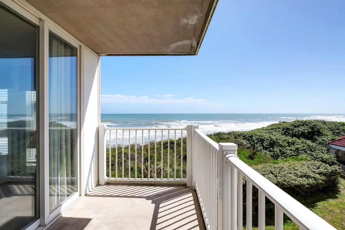 A tranquil balcony with a sturdy railing overlooks the vast ocean, framed by lush greenery below. The inviting space is perfect for relaxing, enjoying a good book, or simply soaking in the sunrise. The gentle curve of the shoreline and the soft ocean horizon create a peaceful backdrop, ideal for couples seeking morning serenity or quiet evening conversations.